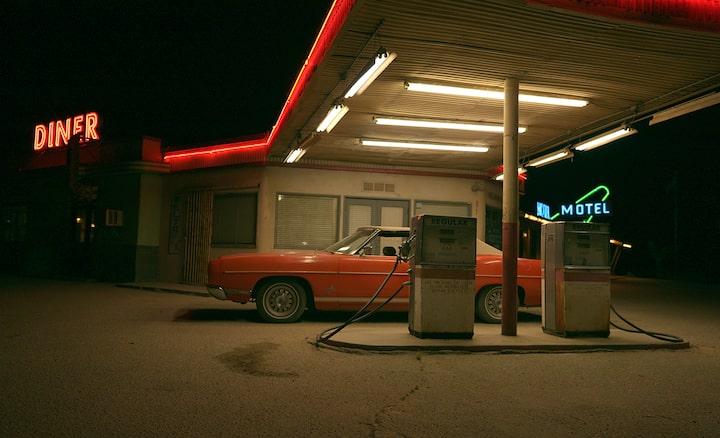 Example image of a vintage red car parked at an old gas station at night, next to an illuminated neon diner sign.